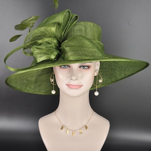 May include: A wide-brimmed olive green hat made of stiffened mesh fabric, featuring a large, structured bow on the side and decorative green leaves. The hat is displayed on a mannequin wearing gold hoop earrings with white pearls and a gold necklace.