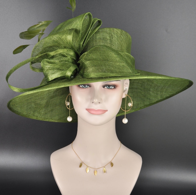 May include: A wide-brimmed olive green hat made of stiffened mesh fabric, featuring a large, structured bow on the side and decorative green leaves. The hat is displayed on a mannequin wearing gold hoop earrings with white pearls and a gold necklace.