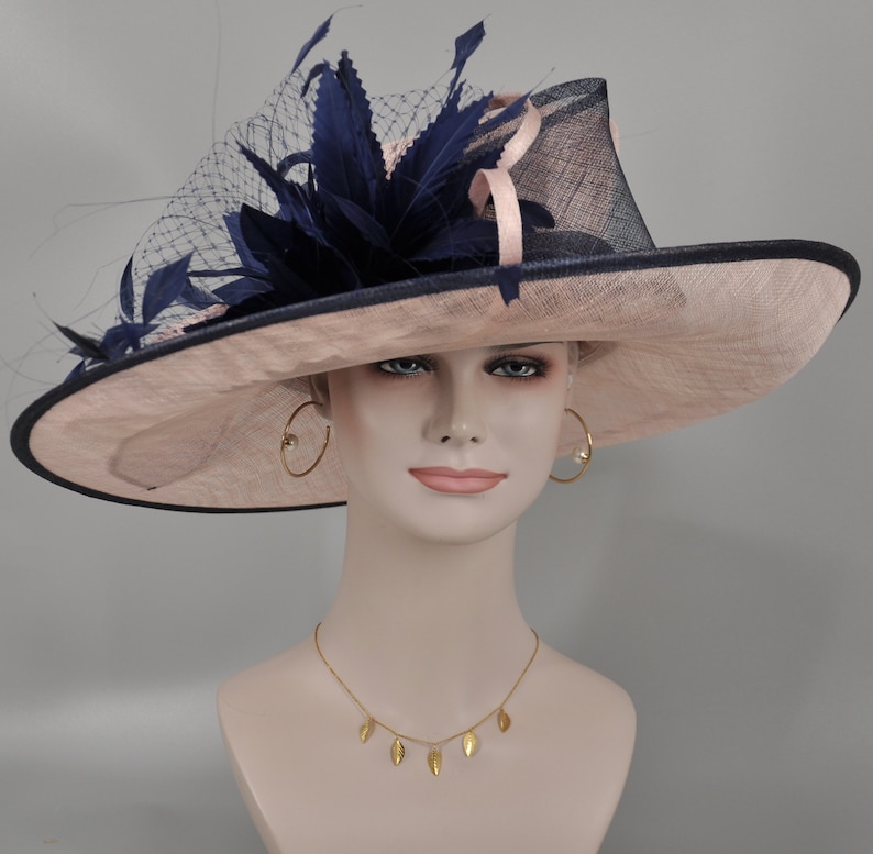 Pu&ograve; includere: Un cappello a tesa larga con base in paglia rosa e bordatura blu navy. Il cappello &egrave; decorato con una grande piuma blu navy e una rete.