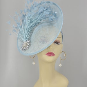 May include: A light blue fascinator hat with a large, round brim and a single, long, light blue feather. The hat is decorated with a silver and crystal brooch.