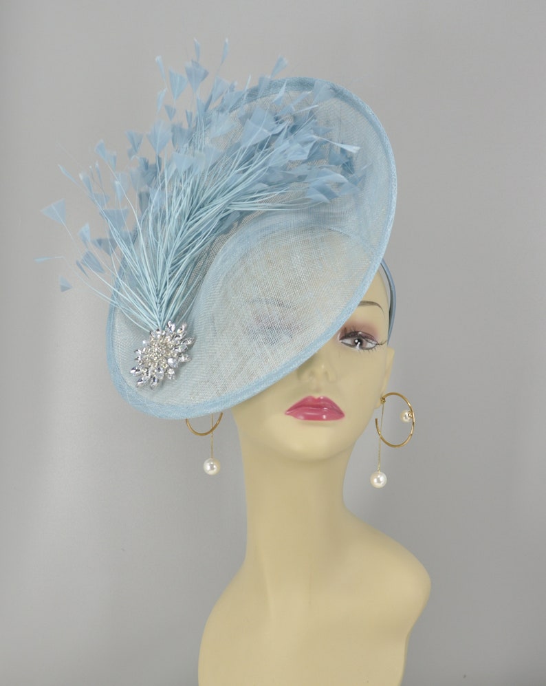 May include: A light blue fascinator hat with a large, round brim and a single, long, light blue feather. The hat is decorated with a silver and crystal brooch.