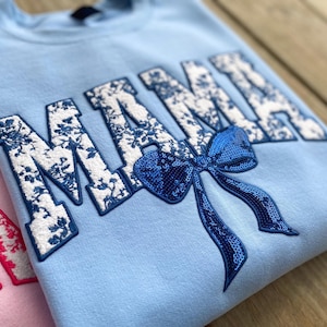 Può includere: Felpa blu chiaro con la parola "MAMA" ricamata in bianco e blu floreale. Un grande fiocco blu di paillettes è centrato sotto la parola.