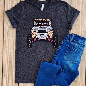 May include: A black t-shirt with a leopard print pattern featuring an embroidered patch of a black and white off-road vehicle with pink accents.