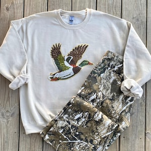 May include: A beige sweatshirt with a green, brown, and white embroidered duck design. The sweatshirt is paired with camouflage pants.