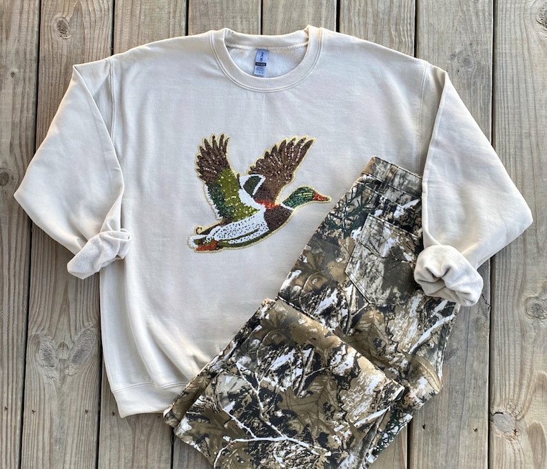 May include: A beige sweatshirt with a green, brown, and white embroidered duck design. The sweatshirt is paired with camouflage pants.