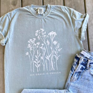 May include: A pale green t-shirt featuring a white floral print and the words "HIS GRACE IS ENOUGH." The shirt is styled with acid wash denim jeans. The t-shirt has rolled sleeves and a round neck.