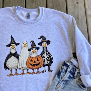 May include: Light gray sweatshirt featuring a Halloween-themed graphic. The design includes four cartoon geese dressed in costumes: a witch, a ghost, a pumpkin, and a skeleton. The sweatshirt is paired with blue jeans.
