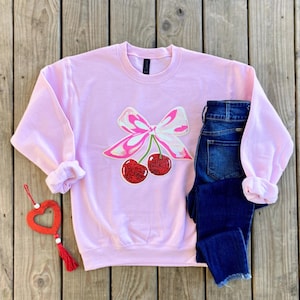 May include: A pink sweatshirt with a cherry and bow design. The cherries are red with a sparkly finish and the bow is pink and white.