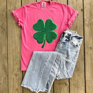May include: A pink t-shirt with a green four-leaf clover made of sequins. The clover is centered on the shirt. The shirt is paired with light blue denim jeans.