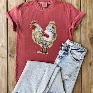 Puede incluir: Una camiseta roja con un gráfico detallado de un gallo, combinada con unos vaqueros de mezclilla de lavado claro. El diseño del gallo presenta acentos rojos, blancos y dorados. Los vaqueros tienen detalles desgastados.
