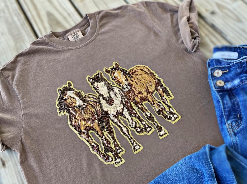 以下が含まれることがあります： 3頭の走る馬の刺繍デザインが施された茶色のTシャツ。馬は茶色、タン、白のアクセントで詳細に描かれています。背景には青いデニムのジーンズが見えます。
