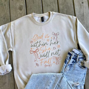 May include: Cream-colored sweatshirt with the text "God is within her, She will not Fall" in orange and brown script. The design includes bow illustrations and leaf accents. Light blue jeans are folded next to the sweatshirt.