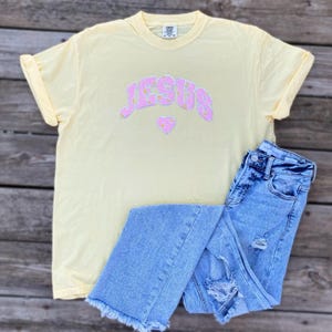 Könnte beinhalten: Ein gelbes T-Shirt mit einem pinkfarbenen "JESUS"-Aufdruck mit kleinen weißen Blumen in den Buchstaben und einem pinkfarbenen Herz darunter. Das Shirt wird mit hellblauen Jeans kombiniert.
