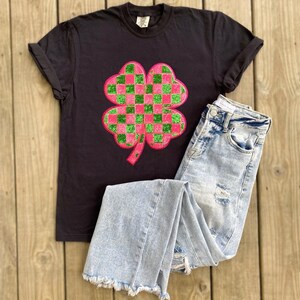 May include: Black t-shirt with a pink and green checkered four-leaf clover graphic.