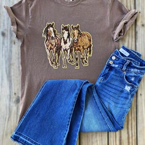 以下が含まれることがあります： 茶色のTシャツ。茶色、ベージュ、白の3頭の馬のグラフィックが特徴です。シャツは青いデニムジーンズと組み合わせています。ジーンズはボタンフライとダメージ加工が施されています。