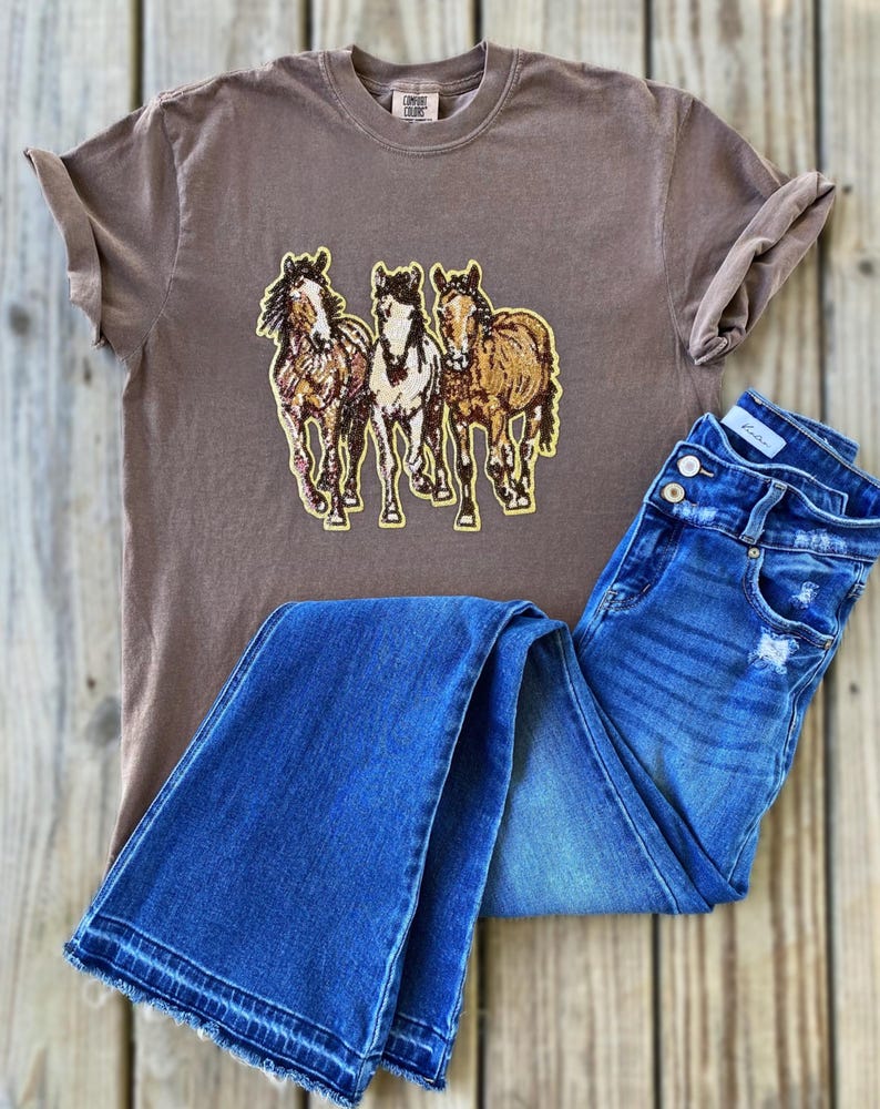 以下が含まれることがあります： 茶色のTシャツ。茶色、ベージュ、白の3頭の馬のグラフィックが特徴です。シャツは青いデニムジーンズと組み合わせています。ジーンズはボタンフライとダメージ加工が施されています。