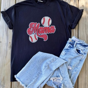 May include: A navy blue t-shirt with the word "Mama" in red script and two baseballs. Light blue acid wash jeans with distressed details are folded next to the shirt. The shirt has rolled-up sleeves.