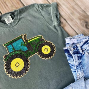 Könnte beinhalten: Ein salbeigrünes T-Shirt mit einem aufgenähten Traktormotiv in Grün, Gelb und Schwarz. Das Shirt wird mit hellblauen Jeans kombiniert. Das Shirt hat ein Comfort Colors-Etikett.