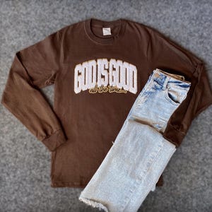 May include: Brown long-sleeved t-shirt with the text "God is good all the time" in white and gold embroidery. The shirt is paired with light blue denim jeans.