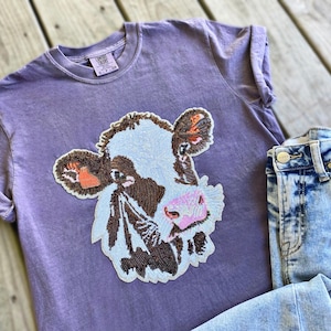 May include: A purple t-shirt with a beaded cow face design and a pair of acid wash denim jeans. The cow face features brown, white, and pink details. The jeans are light blue with a distressed look.