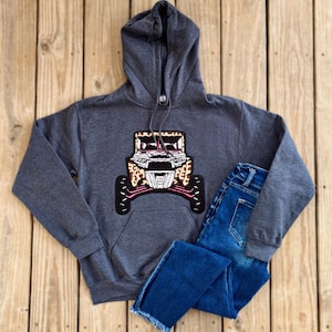 May include: A dark gray hooded sweatshirt with a pink, brown, and white leopard print design of an off-road vehicle on the front.