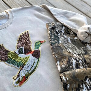 May include: A beige sweatshirt with a sequined duck design. The duck is flying and has green, brown, and white sequins. The sweatshirt is paired with camouflage pants.