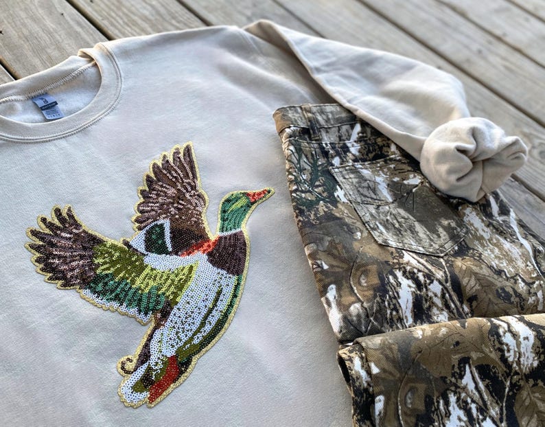 May include: A beige sweatshirt with a sequined duck design. The duck is flying and has green, brown, and white sequins. The sweatshirt is paired with camouflage pants.