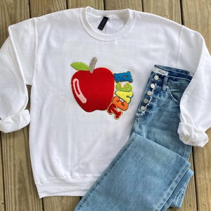 Op de afbeelding: Witte trui met een rode appel en het woord "Teacher" in regenboogkleuren. De trui wordt gecombineerd met lichtblauwe jeans.