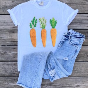 Könnte beinhalten: Weißes T-Shirt mit drei gestickten Karotten auf der Vorderseite. Die Karotten sind orange mit grünen Blättern. Das T-Shirt wird mit hellblauen, ausgefransten Jeans kombiniert.