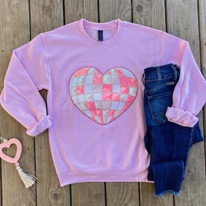 以下が含まれることがあります： ピンクのトレーナーに、大きな虹色のハート型のデザインが施されています。ハートは、ピンク、白、青の小さな正方形でできています。
