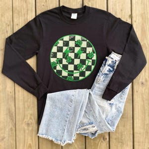 May include: Black long-sleeve shirt with a green and white checkered smiley face graphic. The graphic has green shamrocks around the outside of the smiley face.