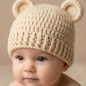 May include: A beige crochet beanie featuring two small, rounded bear ears on top. The hat is crafted with a textured, ribbed stitch pattern that covers the crown and includes a thick, folded brim that sits snugly around the head.