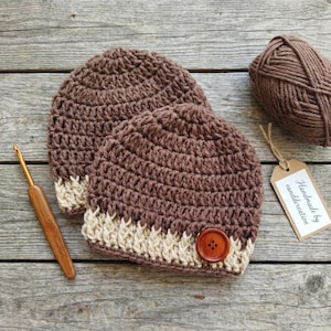 Op de afbeelding: Twee bruine gehaakte mutsen met een crèmekleurige rand en een houten knoop. Een bol bruin garen, een haaknaald en een label met de tekst "Handmade by canddcreation" staan ook op de foto. De mutsen zijn handgemaakt.