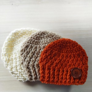 Könnte beinhalten: Drei gehäkelte Mützen in verschiedenen Farben: weiß, beige und orange. Die orangefarbene Mütze hat einen braunen Knopf.