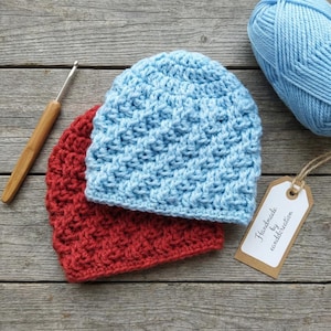 Puede incluir: Dos gorros de ganchillo hechos a mano en rojo y azul claro, con un ganchillo y una madeja de lana azul. Los gorros están hechos con un punto texturizado. Una etiqueta dice "Handmade by canddcreation".