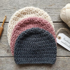 May include: Three handmade crochet beanies in cream, dusty rose, and charcoal gray are stacked on a wooden surface. A wooden crochet hook and a ball of cream-colored yarn are also present. A tag reads "Handmade by canddcreation."