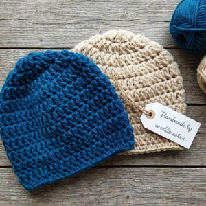 May include: Two handmade crochet beanies in blue and beige, with a crochet hook and yarn balls. The blue beanie is on the left, and the beige beanie is on the right. A tag reads "Handmade by eanddercreation".