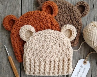 May include: Three handmade crochet bear hats in shades of beige, orange, and brown are displayed on a wooden surface. A crochet hook and a ball of yarn are also present. The hats have bear ears and a textured stitch pattern. A tag reads "Handmade by eanddcreation."