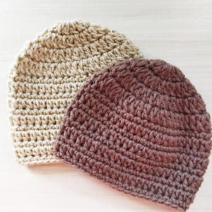 Może przedstawiać: Dwie ręcznie robione czapki z szydełka. Jedna jest w kolorze jasnobeżowym, a druga w kolorze ciemnobrązowym. Czapki wykonane są ściegiem o fakturze i pokazane na jasnej drewnianej powierzchni. Czapki są prawdopodobnie dla dorosłych.