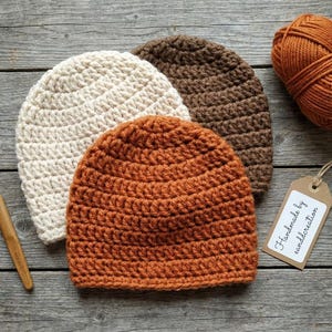 Könnte beinhalten: Drei gehäkelte Mützen in Creme, Braun und Orange, mit einem Knäuel orangefarbener Wolle und einem Häkelhaken. Ein Etikett lautet "Handmade by canddcreation". Die Hüte sind handgefertigt und liegen auf einer verwitterten Holzoberfläche.