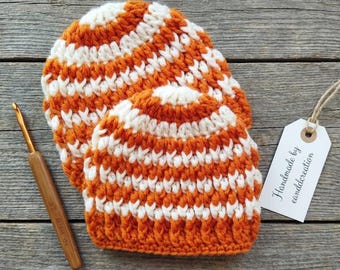 May include: Two orange and white striped crochet hats are displayed on a wooden surface. A crochet hook with a wooden handle rests beside the hats. A tag with the text "Handmade by canddcreation" is attached to the hats.
