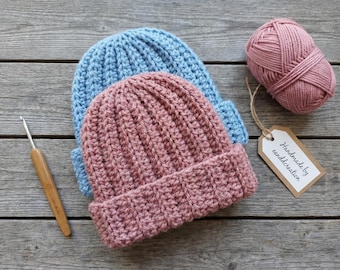 May include: Two handmade crochet beanies, one light blue and one dusty rose, are stacked on a wooden surface. A ball of matching dusty rose yarn and a crochet hook are also present. A tag reads "Handmade by canddcreation."