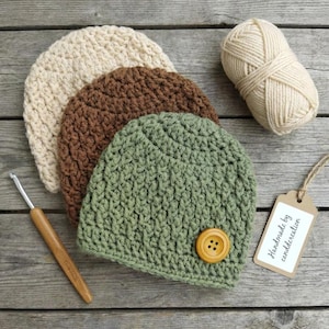 Peut inclure: Trois bonnets crochetés en crème, marron et vert sauge, avec un gros bouton jaune moutarde sur le bonnet vert. Une pelote de laine crème, un crochet en bois et une étiquette en papier marron avec le texte Handmade by canddcreation sont inclus.