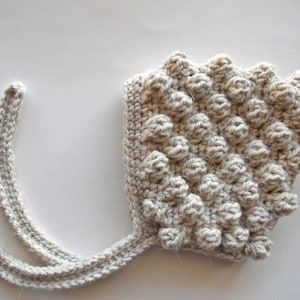 May include: A crocheted baby bonnet with a textured, bobble stitch pattern in a light beige color. The bonnet has a long tie that can be tied under the chin.