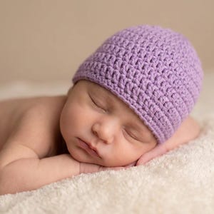 Puede incluir: Un bebé recién nacido durmiendo con un gorro de ganchillo lavanda, descansando sobre una suave manta blanca. El bebé tiene los ojos cerrados y una expresión tranquila. El gorro está hecho a mano con un punto texturizado.