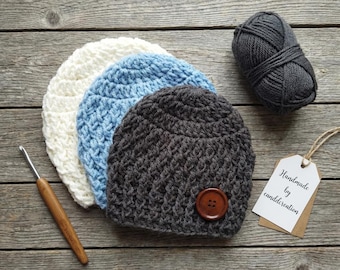 May include: Three handmade crochet beanies in white, light blue, and dark grey, with a wooden button detail on the grey beanie. A ball of grey yarn, a crochet hook, and a tag that says "Handmade by canddcreation" are also present.