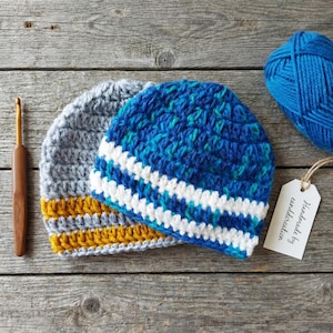 Op de afbeelding: Twee handgemaakte gehaakte mutsen in blauw, grijs en geel. De ene muts is voornamelijk blauw met witte strepen, de andere is grijs met gele strepen. Een bol blauw garen en een haaknaald zijn ook aanwezig.
