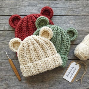 May include: Three crocheted beanies with rounded ears, featuring one dark red, one sage green, and one cream-colored hat. Each hat has a ribbed texture and a folded brim. A wooden crochet hook and a ball of cream yarn are placed nearby.