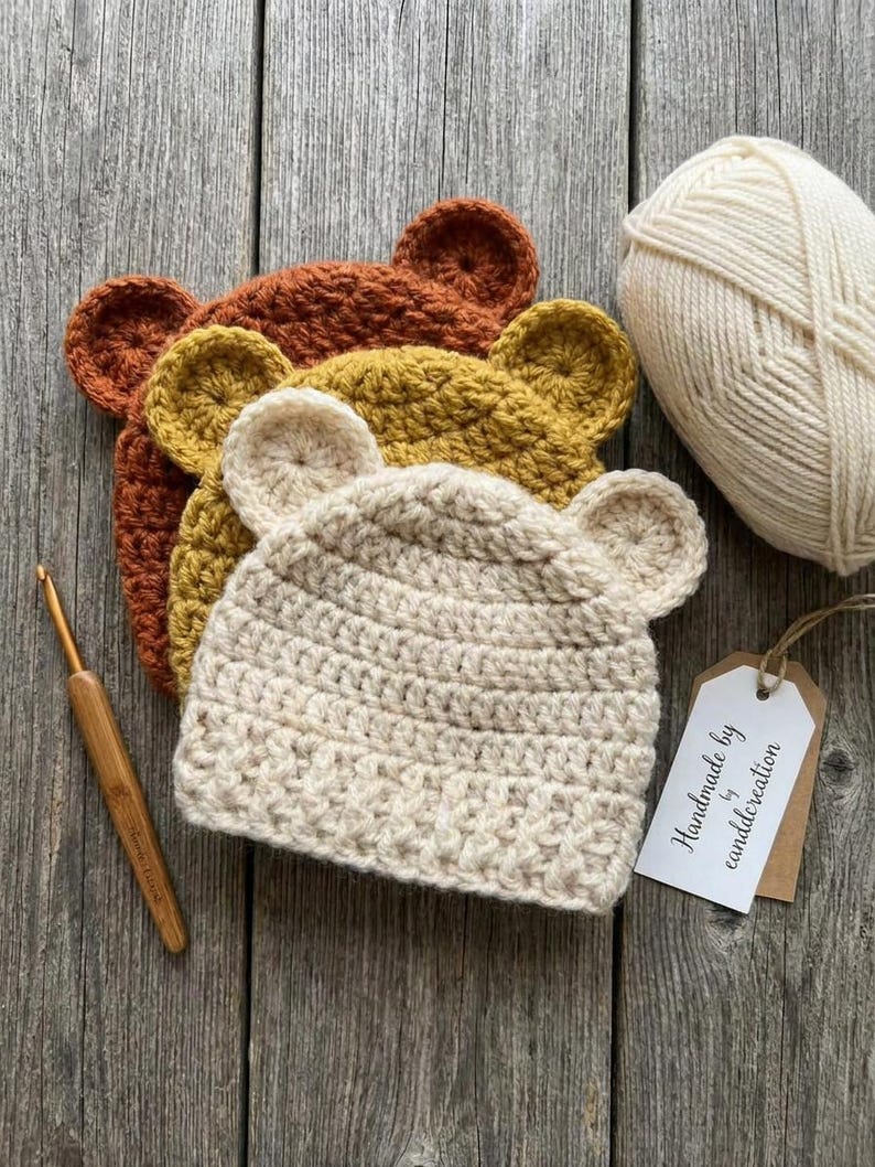 May include: Three crocheted beanies with bear ears in cream, mustard yellow, and rust orange. A ball of cream yarn, a bamboo crochet hook, and a white tag with the text "Handmade by eanddcreation" are arranged alongside the hats.