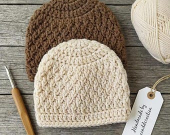May include: Two crocheted beanies, one in dark brown and one in cream, are displayed alongside a ball of cream yarn, a wooden crochet hook, and a white rectangular tag with the text "Handmade by candcreation" printed in black ink.